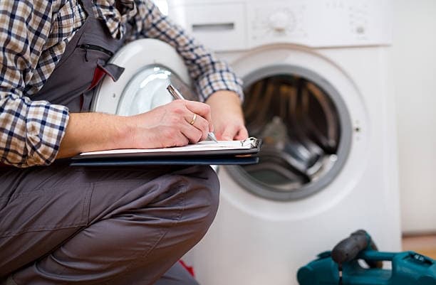 Repairman in grey overalls writing on a clipboard while inspecting an open white washing machine.