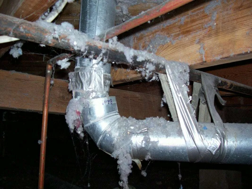 Metal ductwork and pipes in a crawl space heavily covered in thick grey lint.