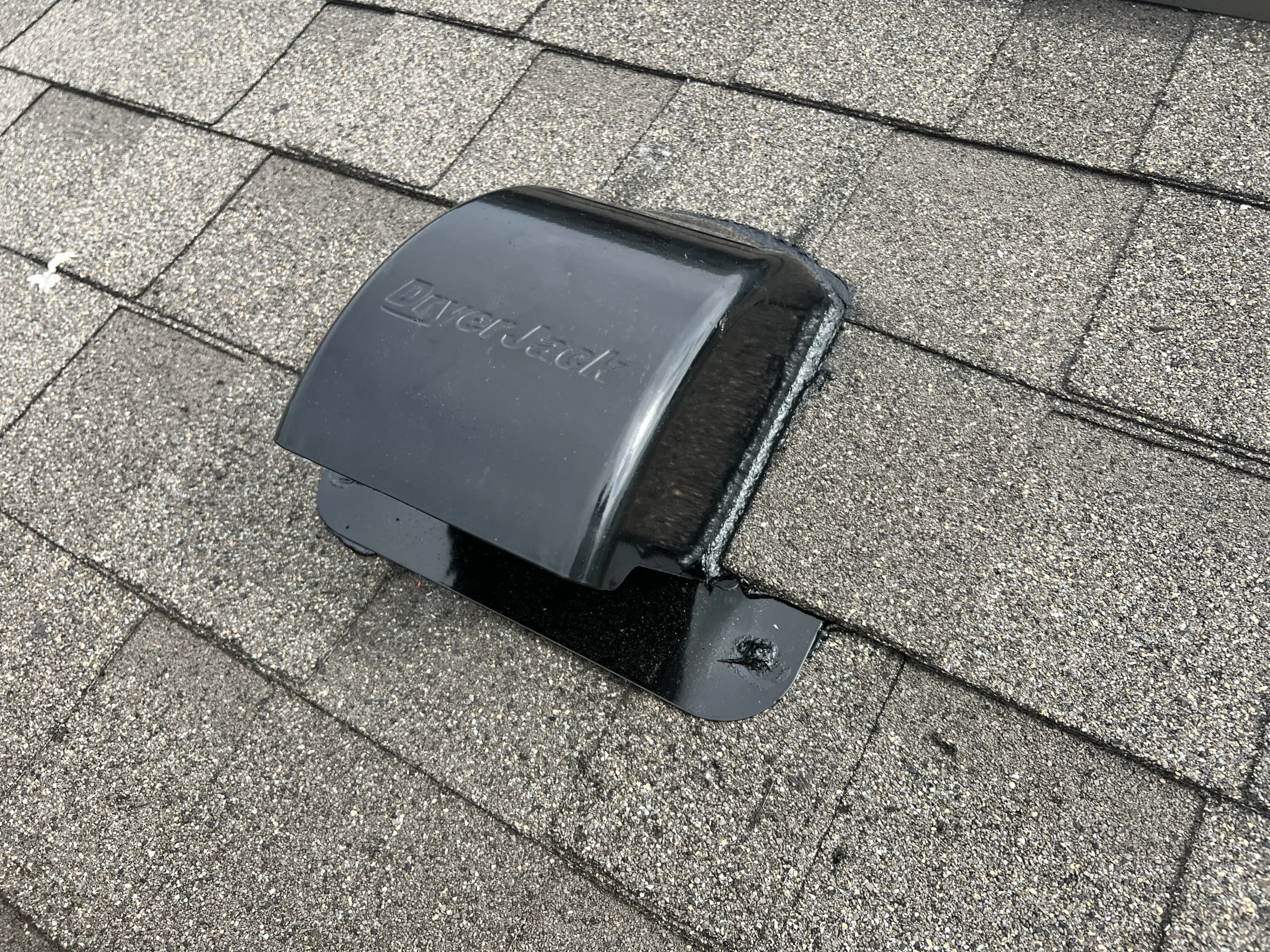 Black DryerJack roof vent installed on grey asphalt shingles for residential dryer exhaust ventilation.