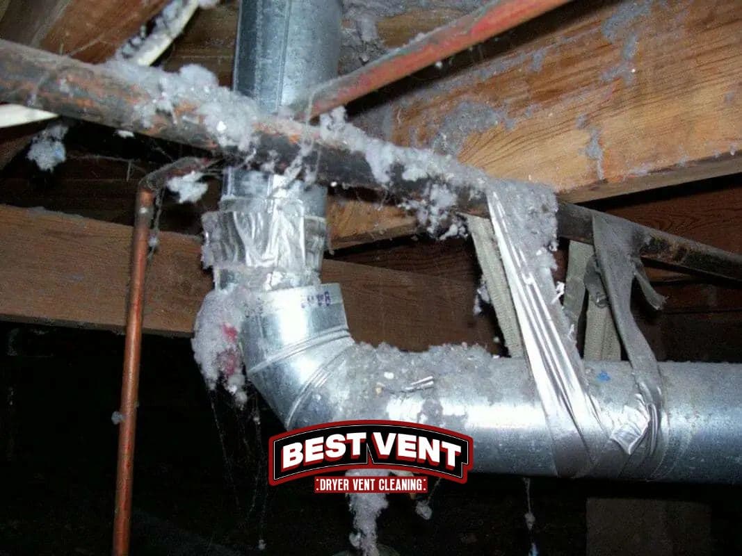 Dirty metal dryer vent pipe heavily covered in grey lint and duct tape in crawlspace.