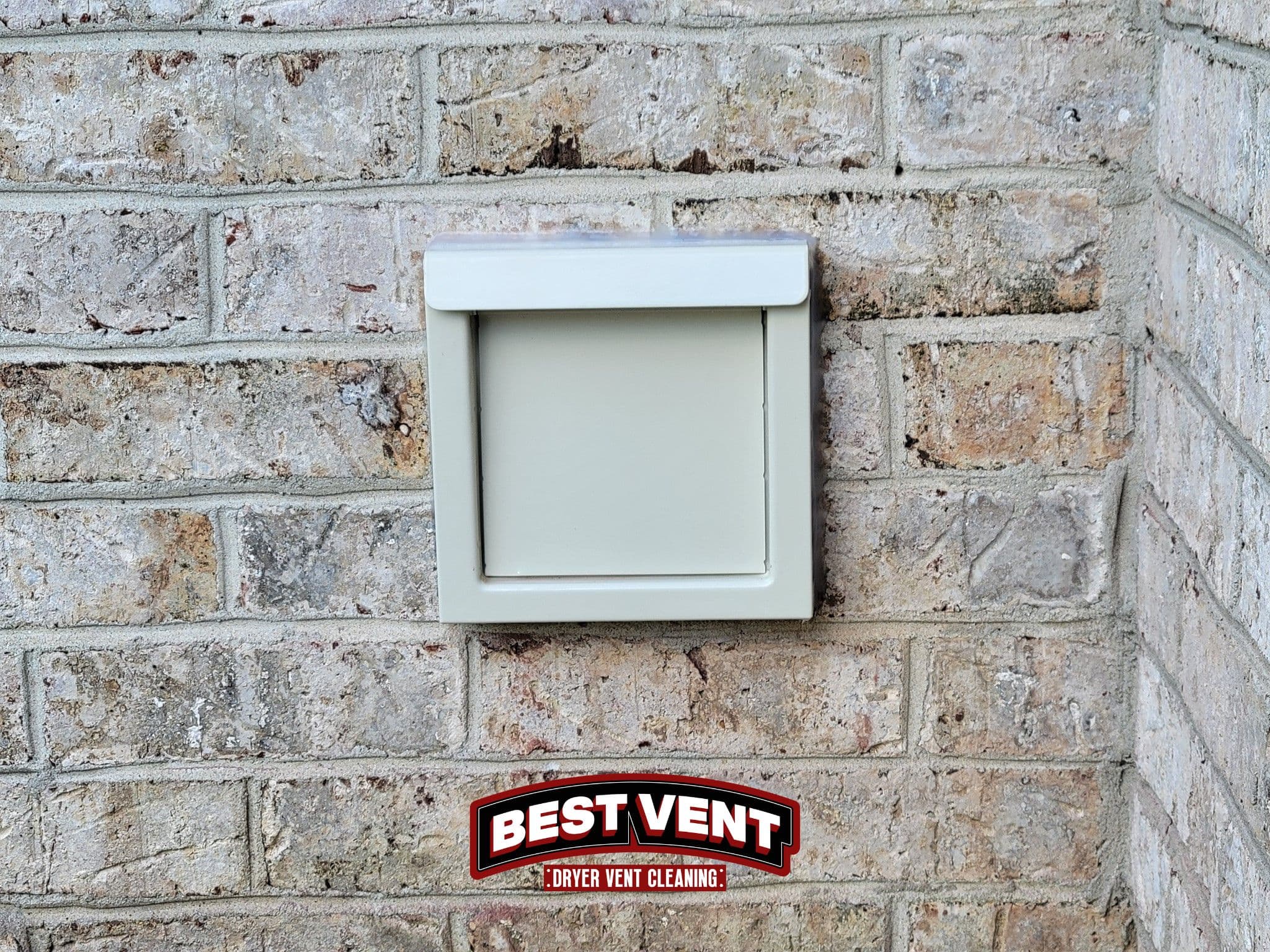 Square tan dryer vent cover with a flap mounted on a light textured brick wall.