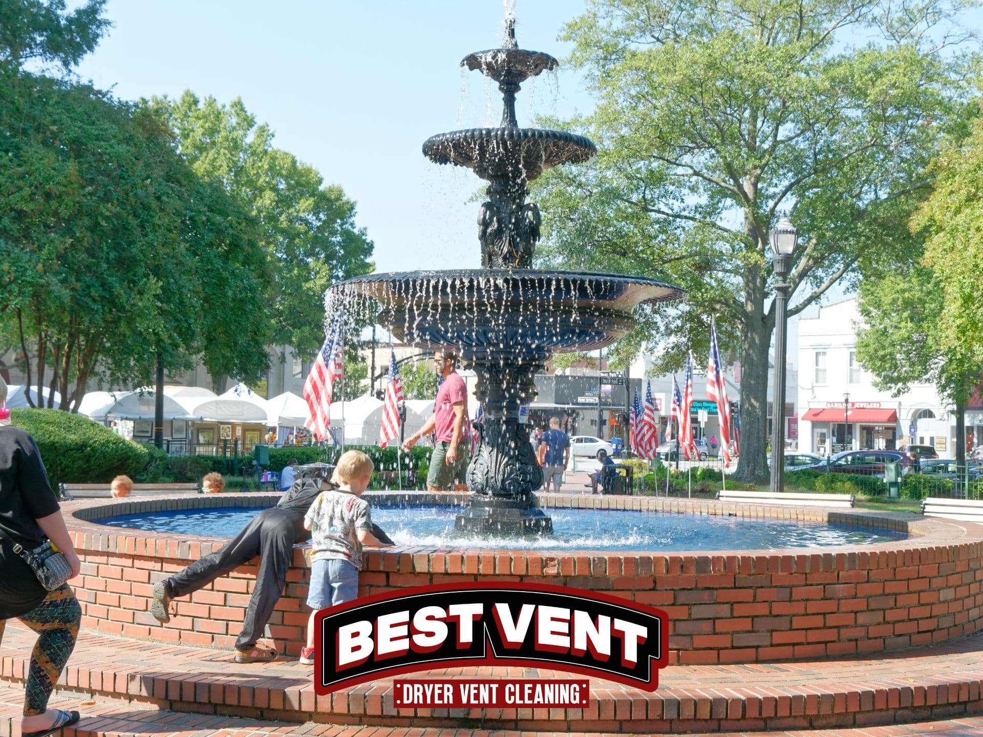 Children playing near an ornate black fountain in a sunny park with American flags.