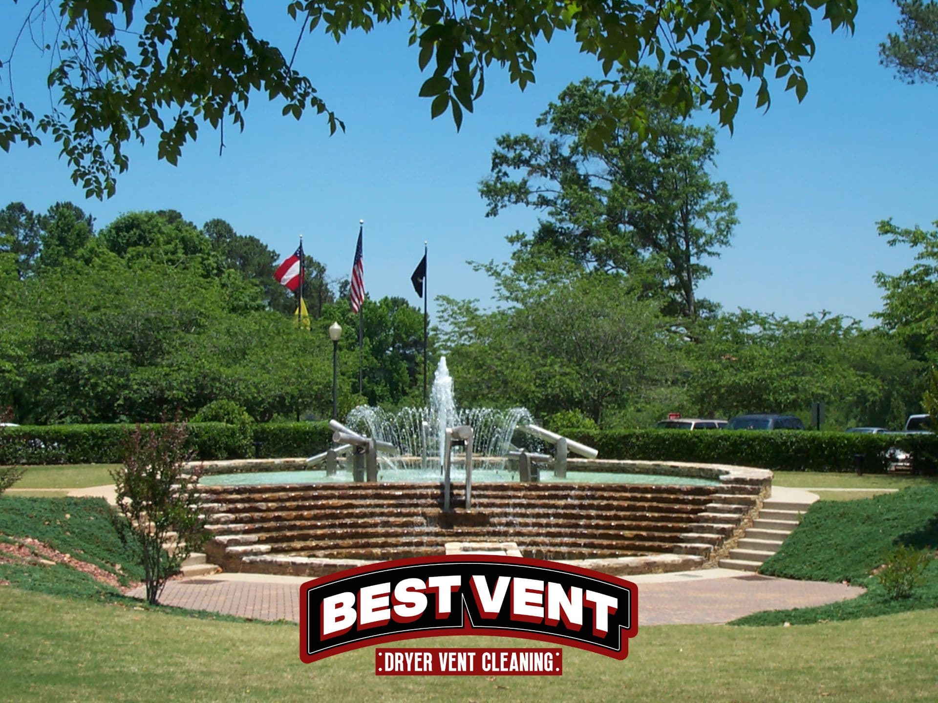 Tiered stone fountain spraying water in a park with flags and Best Vent logo.