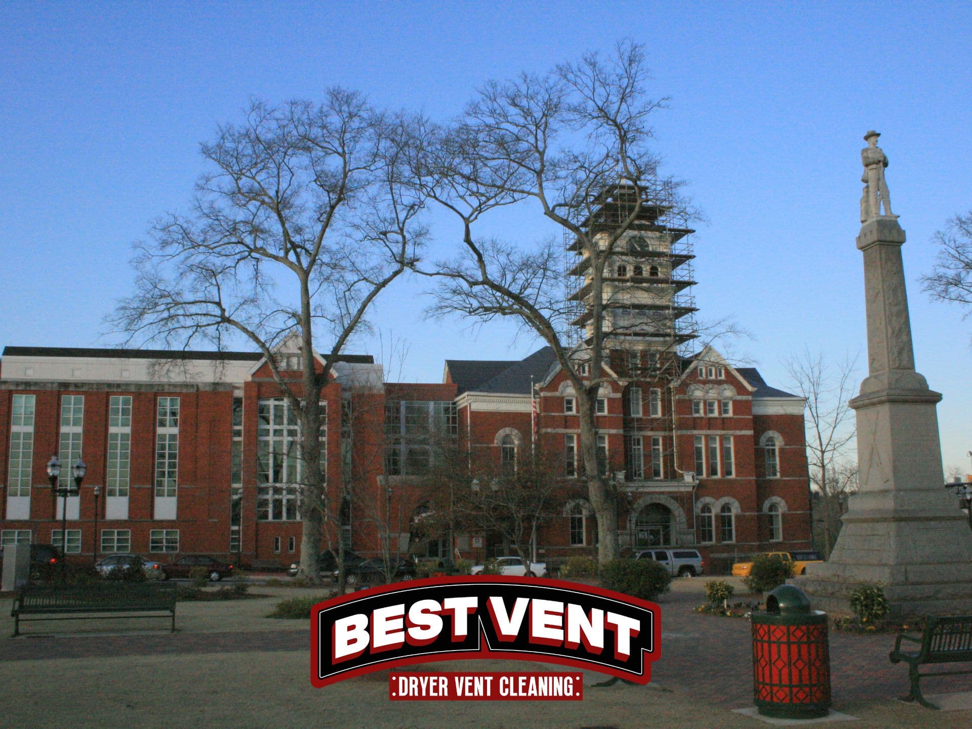Red brick courthouse with a scaffolded clock tower, bare trees, and a tall statue monument.