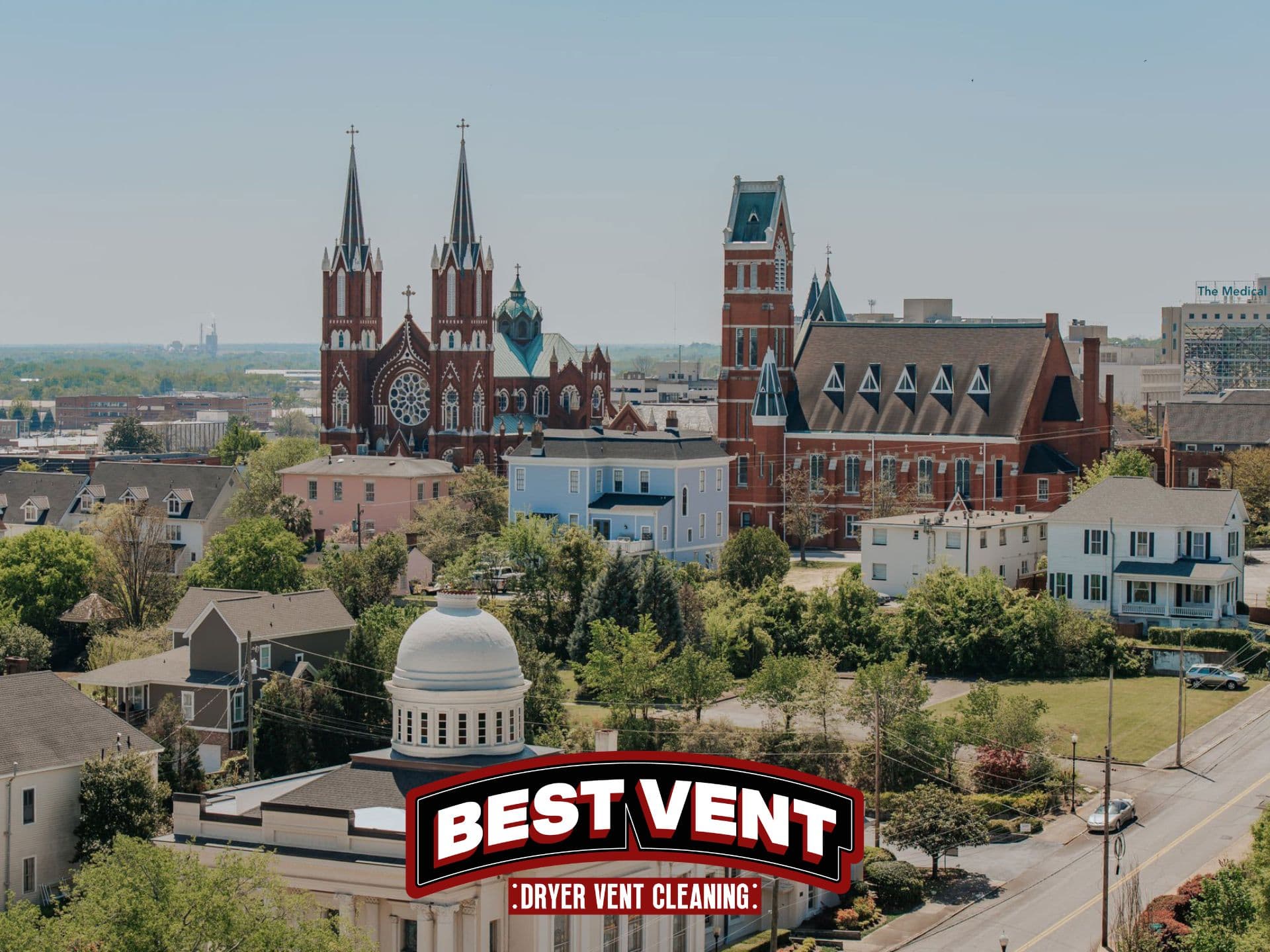Historic cityscape with a twin-spired red brick cathedral, colorful houses, and lush green trees.