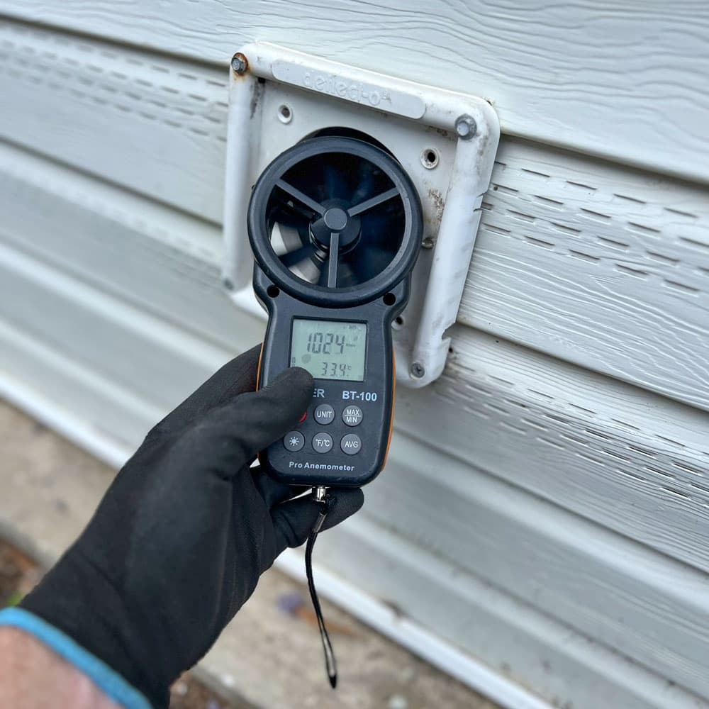 Gloved hand measuring airflow from an exterior wall vent using a digital anemometer.
